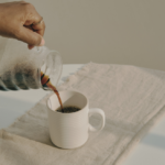 Hand pouring tea from a glass teapot into a white cup on a linen cloth