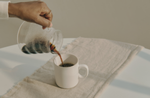Hand pouring tea from a glass teapot into a white cup on a linen cloth