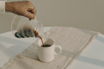 Hand pouring tea from a glass teapot into a white cup on a linen cloth