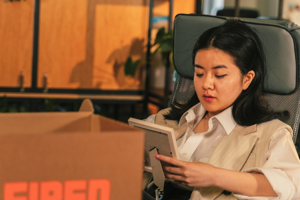 A woman looking slightly concerned sits in an office chair, looking down at a framed photo. A cardboard box sits on the foreground suggesting packing or unpacking from her office desk